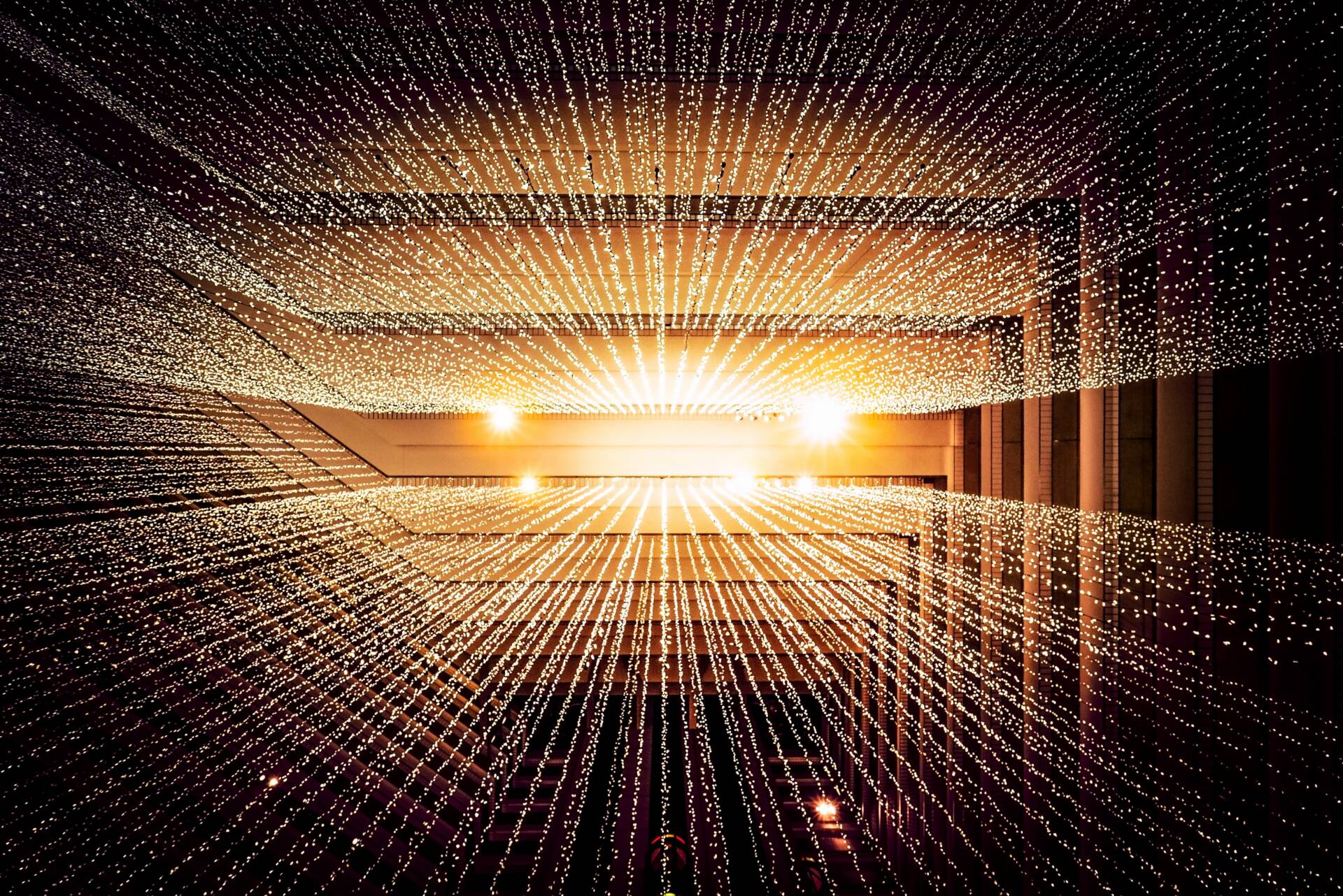 Photo looking upward through cascading light strings inside a square building with a spiraling, open-center design.