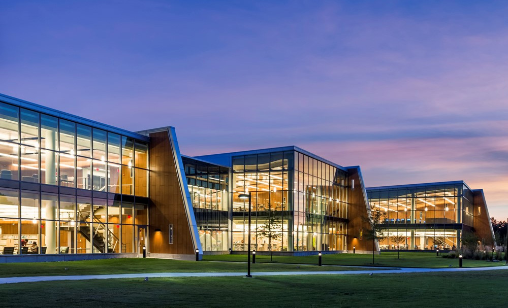 Exterior view of a large glass building at night with angled walls lined in wood paneling, lights on, and students visible inside.